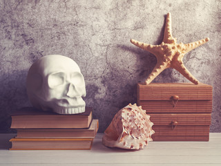 A decorative figure in the form of a human skull is on a pile of books. Nearby is a wooden box on...