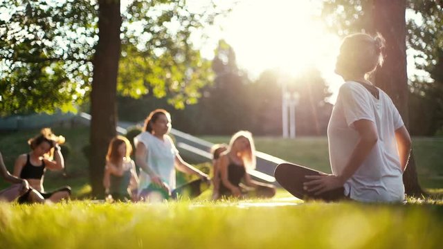 Group of women is stretching necks in park on summer sunny morning under guidance of coach in slow motion. Group of people outdoors is sitting on yoga mat in lotus pose on grass with their eyes closed