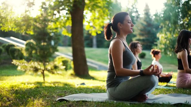 Young woman sitting on yoga mat in lotus pose and meditating in a park at sunrise. Girl opens her eyes and starts to rotate her shoulders. Tracking shot in slow motion