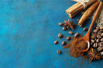 coffee grains scattered on a blue textural background, anise stars, cinnamon sticks and ground coffee in a wooden spoon. Top view