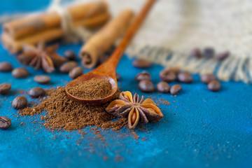 coffee grains scattered on a blue textural background, anise stars, cinnamon sticks and ground coffee in a wooden spoon.