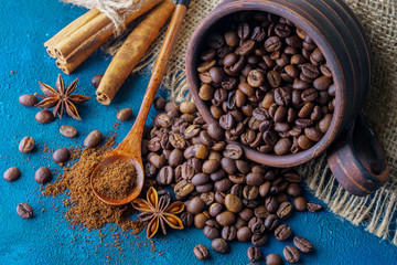 coffee grains pouring out of a clay cup  and scattered on a blue textural background, anise stars, cinnamon sticks and ground coffee in a wooden spoon.