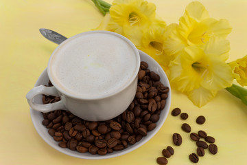 A cup of cappuccino coffee with milk crema and coffee beans on a yellow table, where there is a delicate yellow gladiolus flower. The concept of a soft and gentle summer cappuccino in yellow tones.