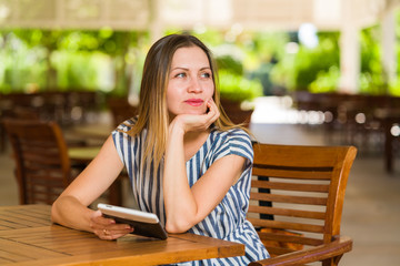 Beautiful business woman thoughtfully looks far in the distance with a smile on her face. She is sitting at the outdoor park terrace at the wooden table while holding tablet or e-ink book in her hand.