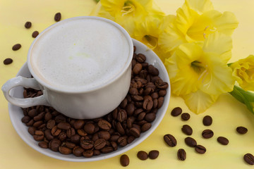 A cup of cappuccino coffee with milk crema and coffee beans on a yellow table, where there is a delicate yellow gladiolus flower. The concept of a soft and gentle summer cappuccino in yellow tones.