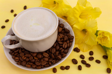A cup of cappuccino coffee with milk crema and coffee beans on a yellow table, where there is a delicate yellow gladiolus flower. The concept of a soft and gentle summer cappuccino in yellow tones.