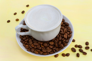 Cup of cappuccino coffee with milk froth and coffee beans on a yellow table, closeup. The concept of a soft and gentle summer cappuccino in yellow tones.