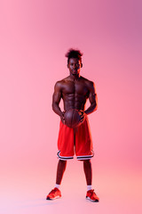 Fototapeta na wymiar confident african american basketball player with muscular torso looking at camera on pink and purple gradient background with lighting