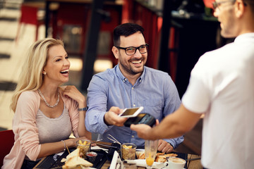 Cheerful guests paying with mobile phone while having lunch in a restaurant.