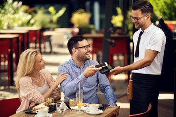 Happy couple paying for lunch via contactless smart phone payment in a restaurant.