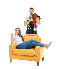 Sad househusband with laundry and his lazy wife sitting in armchair against white background