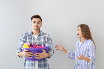 Angry wife scolding her sad husband with cleaning supplies on grey background