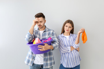 Young wife and stressed husband with cleaning supplies on grey background