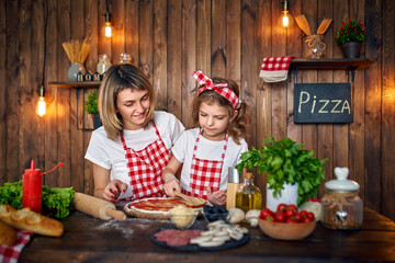 Happy mother and daughter wearing white T-shirts and checkered aprons cooking pizza together and decorating with mushrooms on table filled with ingredients, in stylish wooden kitchen.