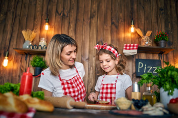 Happy mother and daughter wearing white T-shirts and checkered aprons cooking pizza together and decorating with mushrooms on table filled with ingredients, in stylish wooden kitchen.
