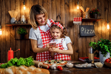 Beautiful mother and daughter wearing white T-shirts and checkered aprons and headbands smiling and take pictures of cooked pizza on pink cellphone, in stylish kitchen interior.
