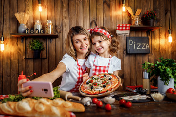 Beautiful mother and daughter wearing white T-shirts and checkered aprons and headbands smiling and taking selfie with cooked pizza on pink cellphone, in stylish kitchen interior.