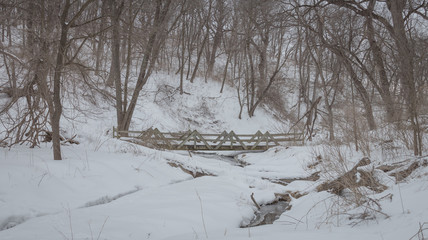 A wooden bridge spans a small creek in a snow covered forest.