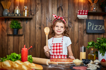 Smiling girl wearing white T-shirt with checkered apron and headband cooking pizza on table filled with ingredients for pizza and looking at camera, in stylish wooden kitchen.