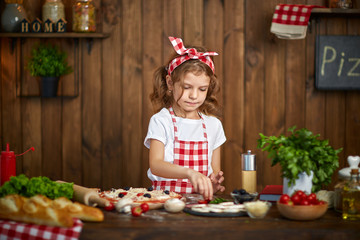 Smiling girl wearing white T-shirt with checkered apron and headband cooking pizza on table filled with ingredients for pizza in stylish wooden kitchen