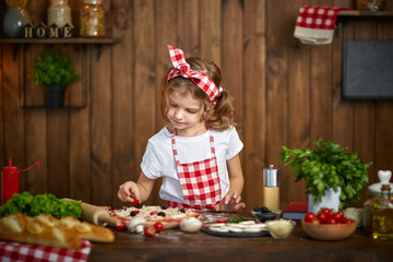 Smiling girl wearing white T-shirt with checkered apron and headband cooking pizza on table filled with ingredients for pizza in stylish wooden kitchen