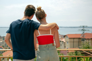 Naklejka premium The young man and the girl, embracing, stand on the balcony and look at the landscape. The photo is taken from the back.