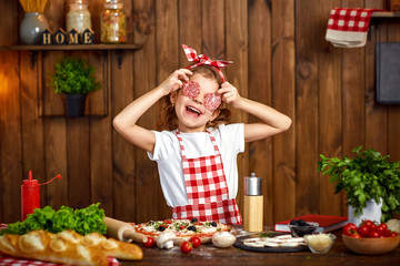 Adorable little female chef wearing checkered apron and headband cooking pizza and making face with...
