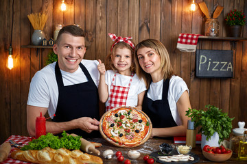 Beautiful family wearing white T-shirts and aprons sitting by table filled with ingredients and showing cooked delicious pizza, looking at camera in stylish wooden kitchen.