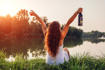 Young woman enjoying glass of wine on river bank at sunset raising arms and feeling free and happy.