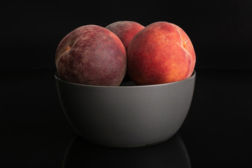 Group of three whole fresh red peach in a gray ceramic bowl isolated on black glass
