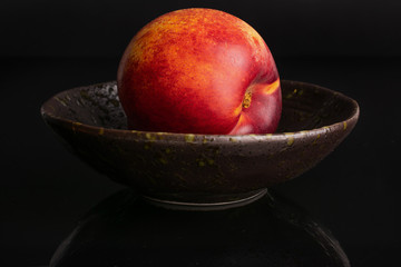 One whole fresh red nectarine in a dark ceramic bowl isolated on black glass