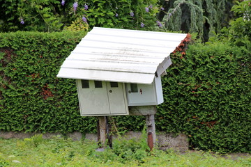 Electronic meter box and fuse box mounted on dilapidated wooden poles and covered with metal sheet protection surrounded with uncut grass and hedge at abandoned construction site on warm sunny summer 