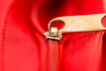 Metal zipper on intense red leather jacket or purse detail close up macro. The zipper is partly open and binding together the two textile layers with thread. Handle on the upper side.