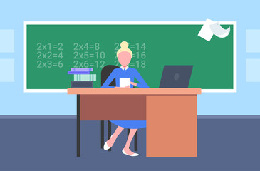 female teacher sitting at desk in front of chalkboard using laptop math lesson education concept modern school classroom interior horizontal flat