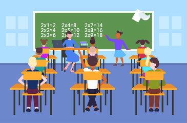 female teacher with pupils sitting at desks looking at schoolgirl solving math problem at chalkboard school lesson education concept modern school classroom interior flat full length horizontal