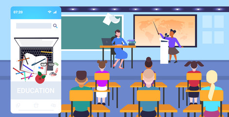 teacher with pupils sitting at desks looking at schoolboy speaking at world map geography lesson school education concept online mobile app classroom interior full length horizontal