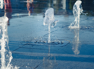 Obraz na płótnie Canvas Water streams flowing out of stainless grate, close up view. Mother with kid reflecting on water surface. Summer square with dry fountain. Selective soft focus. Blurred background