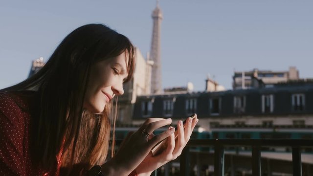 Happy beautiful tourist woman using smartphone social network app at morning cafe balcony with sunny Eiffel Tower view.