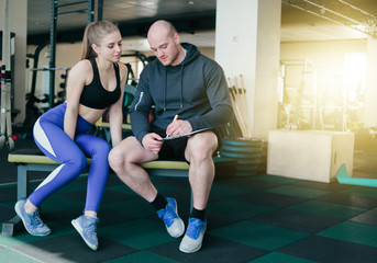 Fototapeta na wymiar The concept of personal training. Instructor notes in notebook results training sporty blonde woman sitting on a bench. Smiling sports couple