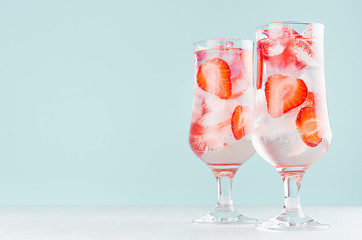 Detox cold water with ripe strawberry slices, ice cubes and tonic in two elegant glasses on white wood table, mint color wall.