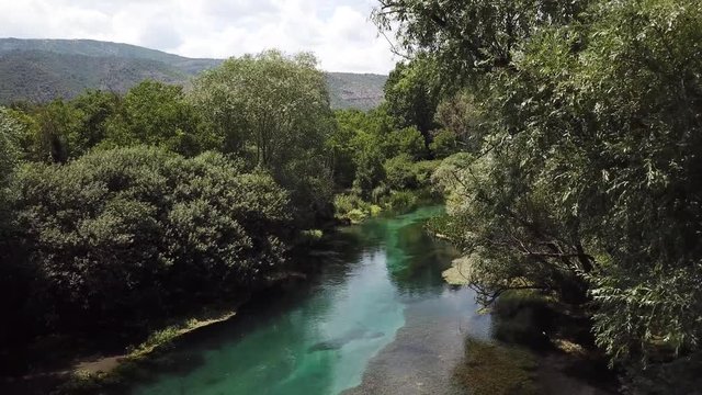 Beautiful slow drone clip of a descent through the trees to just above a turquoise crystal clear river below. The river is the Tirino in the Abruzzo region in Italy. Lush vegetation and Mountains.