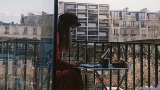 Camera slides right revealing beautiful journalist woman using laptop at idyllic summer Paris apartment balcony.