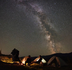 Silhouettes of young family hikers having a rest together in summer camping, sitting on chairs beside campfire and tourist tents at night. Guys enjoying night starry sky full of stars and Milky way