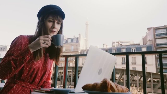 Beautiful elegant businesswoman using laptop, enjoying coffee at idyllic Paris morning balcony with Eiffel tower view.