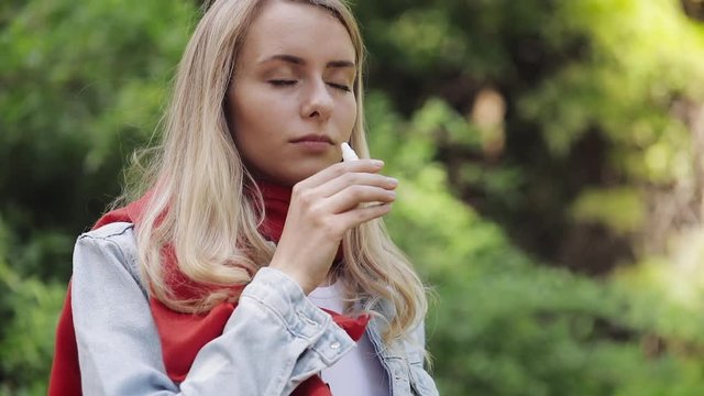 Young Woman Wearing Red Scarf Spraying Nasal Spray Standing in The City Park. Health Care Therapy Concept Concept. Medicine.