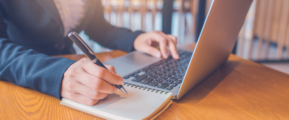 Business women hand are taking notes on paper with a black pen, and she is using a laptop computer on a wooden desk in the office.Web banner.