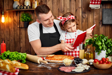 Girl wearing checkered apron reading red recipes book while father spreading sauce by dough for pizza in stylish wooden kitchen