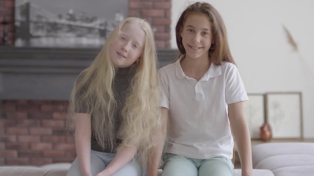 Two cute smiling girls sitting on the couch ooking at camera in the living room. Concept of friendship. Carefree childhood. Adorable girls indoors. Real people series.
