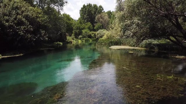 Slow drone clip flying in reverse through the trees just above the crystal clear river Tirino in the Abruzzo region in Italy. A dragonfly can be seen following.