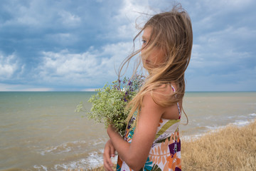 Portrait of a girl with a bouquet of wild flowers against the background of the sea and blue sky with clouds